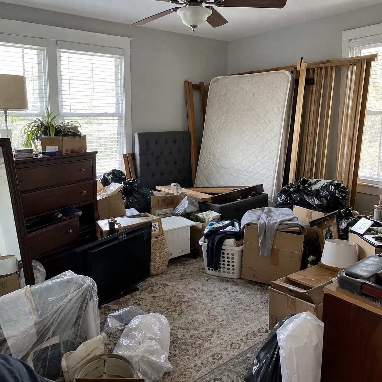 A bedroom stacked to the window with a mattress, two dressers, bagged clothing and moving boxes before a cleanout