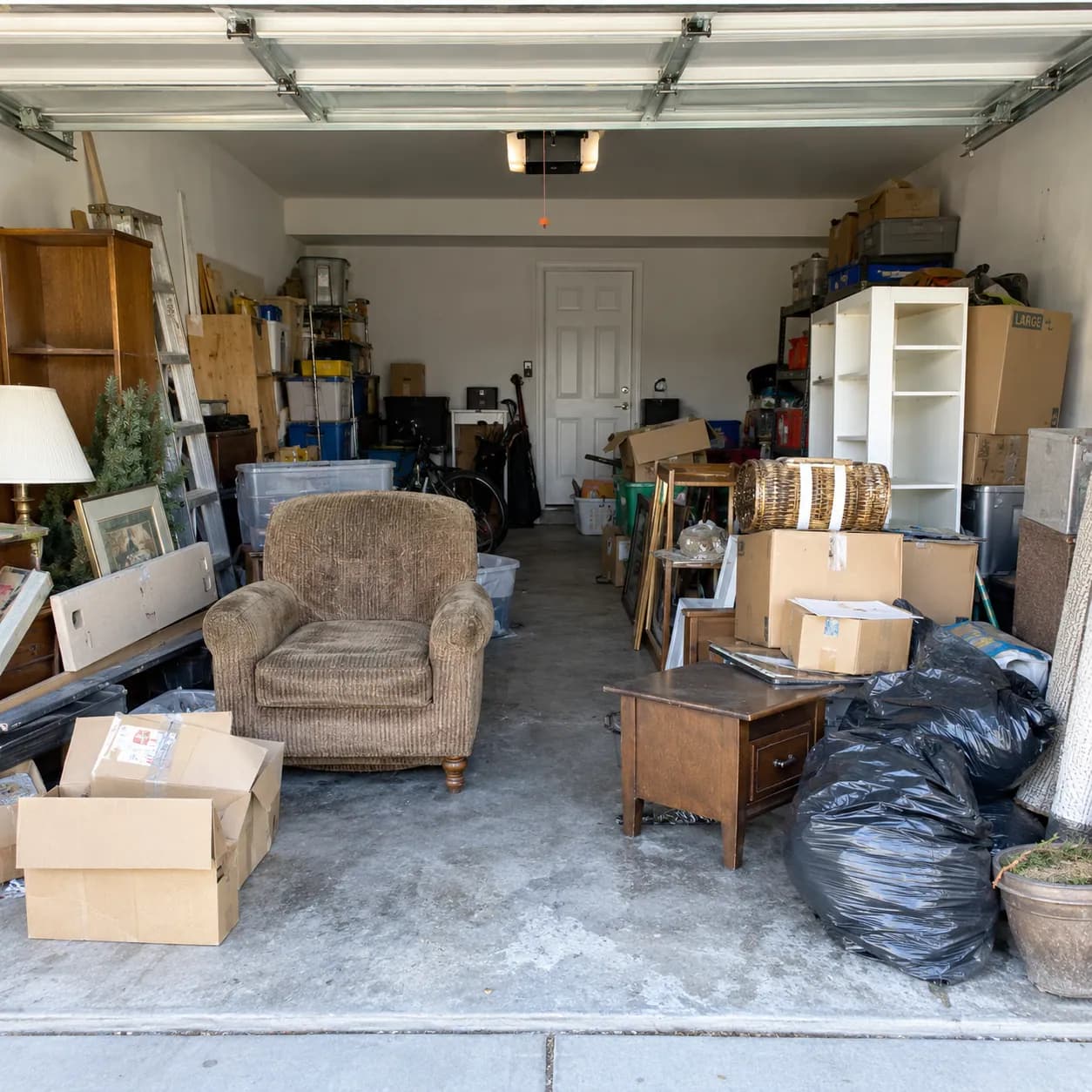 A two-car garage packed wall to wall with boxes, an armchair, shelving and bagged clutter before a cleanout